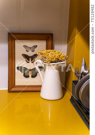 White vase with yellow wildflowers near frame with dried butterflies in kitchen 73128482