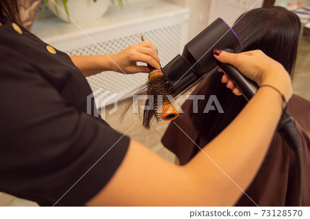 Closeup of young female hands holding a hair dryer and professional round brush and making a hairstyle to brunette customer in beauty salon. 73128570