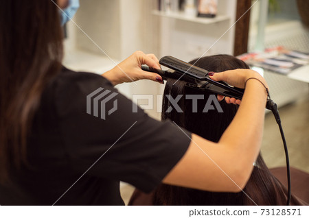 Close up image of unrecognizable hairdresser in black uniform while making a hairstyle using a hair straightener 73128571