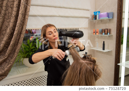 Beautiful adult woman hairdresser making a hairstyle to blonde customer using a hair dryer and round brush. 73128720