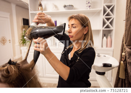Elegant adult woman hairdresser enjoying working in beauty salon and blowdrying hair of customer 73128721