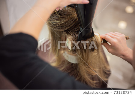 Closeup of hands of hairdresser while blowdrying hair of customer in the hair salon Closeup of hands of hairdresser while blowdrying hair of customer in the hair salon 73128724