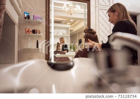 Image on the mirror of young woman getting haircut by adult elegant hairdresser in the beauty salon 73128734