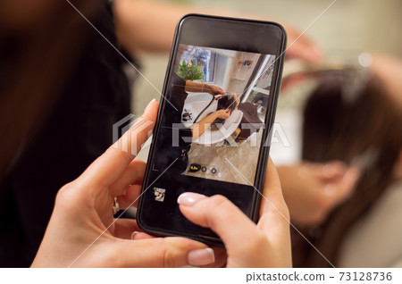 Closeup of manicured hands holding a black smartphone in live view mode and taking a picture of a hair wash in beauty salon 73128736