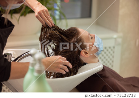 Young woman in protective medical mask getting a hair wash in beauty salon during pandemic. Concept of safety hairdresser work during pandemic 73128738