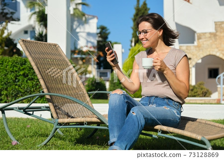 Beautiful woman resting sitting on sunbed on green lawn, reading smartphone 73128869
