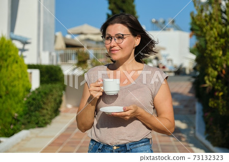 Mature woman in glasses with cup of coffee walking outdoor 73132323