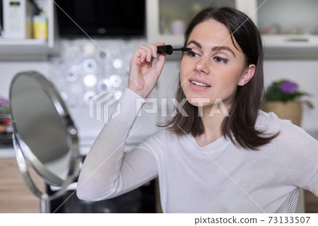 Young woman applies mascara on eyelashes using mirror Young woman applies mascara on eyelashes using mirror 73133507