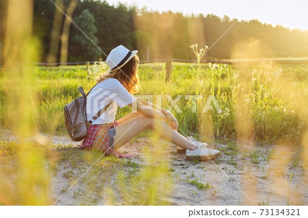 Happy teenager girl in hat with backpack sitting on country road, enjoying summer vacation 73134321