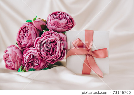 White present box with pink ribbon and violet peonies flowers on white silk fabric background. Greeting card for holidays. White present box with pink ribbon and violet peonies flowers on white silk fabric background. Greeting card for holidays. 73146760