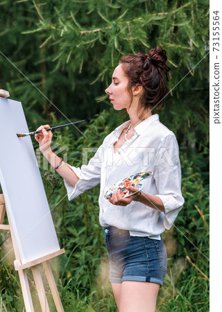 Girl artist, woman paints a picture, summer forest, white shirt denim shorts, creating creativity and artistic mood. First brush stroke, getting started. Background of green grass trees and spruce. 73155564