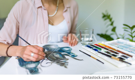Close-up portrait, female hands draw a picture with paints, brushes and color paint palette. Tea service teapot and bottles. Creativity and creation, at home in the studio. Close-up portrait, female hands draw a picture with paints, brushes and color paint palette. Tea service teapot and bottles. Creativity and creation, at home in the studio. 73155770