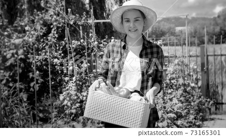 Black and white portrait of smiling female farmer posing with fresh ripe vegetables in box at garden Black and white portrait of smiling female farmer posing with fresh ripe vegetables in box at garden 73165301