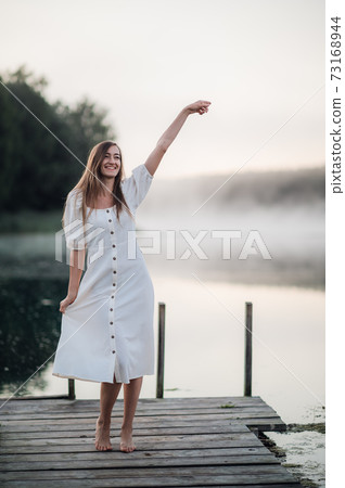 Happy woman in a white dress enjoys morning on a lake. Fog floats over the water. Happy woman in a white dress enjoys morning on a lake. Fog floats over the water. 73168944
