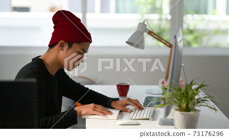 Side view of freelance graphic designer sitting at his workspace and looking at notebook while working on computer. 73176296