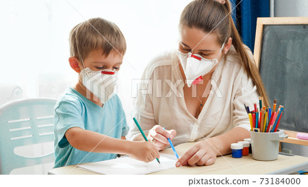 Little boy and young mother wearing medical mask respirator doing homework behind desk at home. Protecting children and families from virus during lockdown and self isolation. Remote school and 73184000