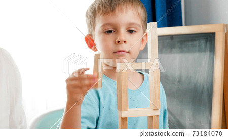 Happy smiling boy putting wooden block on top of high tower made of toy wooden blocks and bricks. Concept of children education at home during lockdown and staying at home 73184070