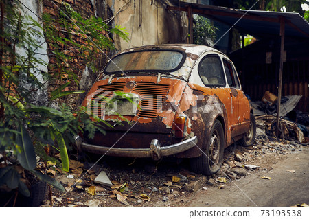 Old abandoned rusty white and orange vintage car wreck in an old street 73193538