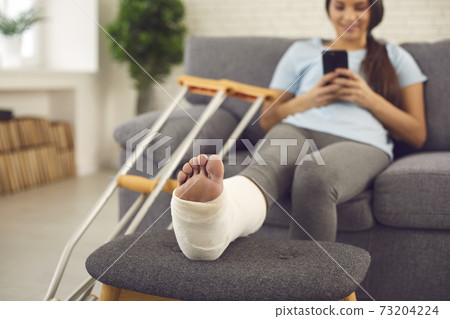 Woman with broken leg in plaster cast sitting on couch at home with mobile in hands 73204224