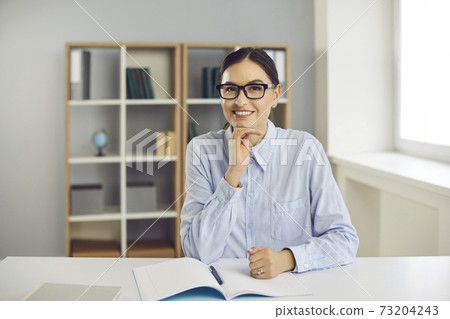 Happy young school teacher or college student sitting at desk and smiling at camera 73204243