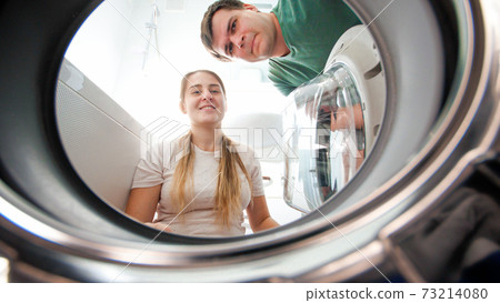 Smiling young couple looking inside washing machine and taking out clothes at laundry. Family doing housework and laundry together Smiling young couple looking inside washing machine and taking out clothes at laundry. Family doing housework and laundry together 73214080