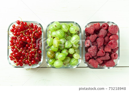 Assorted berries of raspberries, gooseberries and red currants in glass bowls on a white table. 73218948