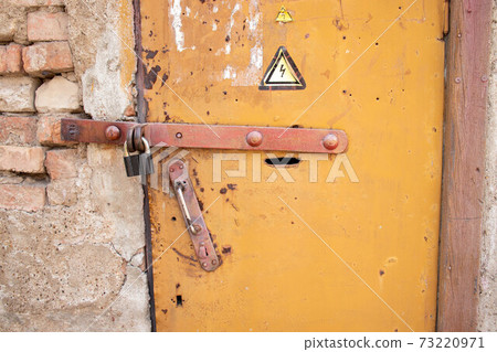 Locked old rusty yellow door with high voltage sign. Grey wall of transformer substation and padlock 73220971