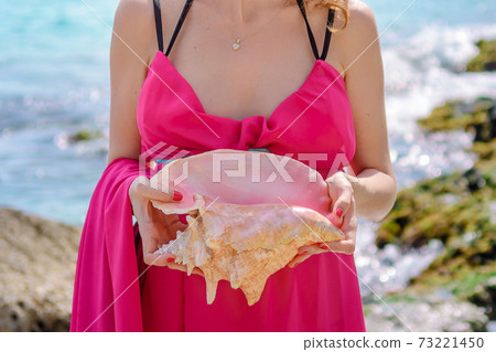 The hands of a pregnant woman in a pink dress on her stomach hold a seashell. 73221450