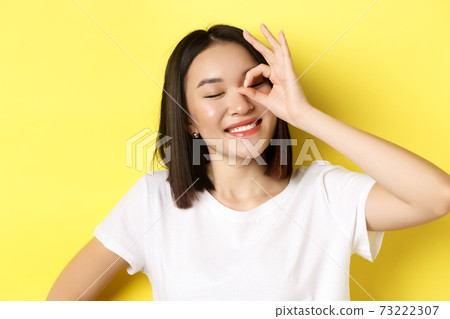 Close up of cute asian girl feeling happy, showing OK sign on eye and smiling, standing over yellow background Close up of cute asian girl feeling happy, showing OK sign on eye and smiling, standing over yellow background 73222307