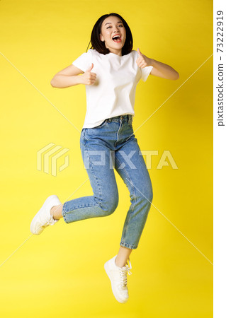 Full size of happy young asian woman jumping from joy, showing thumbs up in approval, posing over yellow background in jeans and casual white t-shirt Full size of happy young asian woman jumping from joy, showing thumbs up in approval, posing over yellow background in jeans and casual white t-shirt 73222919