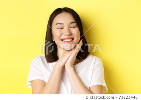 Close up of romantic asian woman dreaming of something cute, close eyes and hold hands near face, imaging beautiful thing, standing over yellow background Close up of romantic asian woman dreaming of something cute, close eyes and hold hands near face, imaging beautiful thing, standing over yellow background 73223446