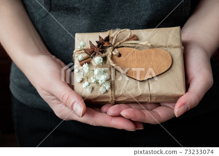 Gift box wrapped in craft paper decorated with wooden heart and white flowers in human hands. Valentine day celebrating Gift box wrapped in craft paper decorated with wooden heart and white flowers in human hands. Valentine day celebrating 73233074