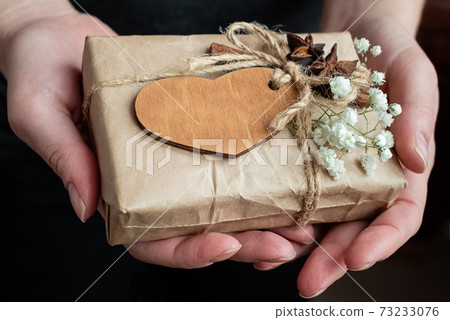 Gift box wrapped in craft paper decorated with wooden heart and white flowers in human hands. Valentine day celebrating Gift box wrapped in craft paper decorated with wooden heart and white flowers in human hands. Valentine day celebrating 73233076