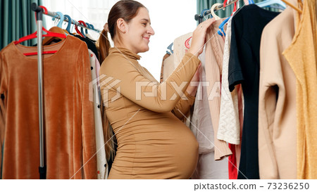Portrait of smiling pregnant woman standing between long clothes rack in shopping store and holding beautiful dress Portrait of smiling pregnant woman standing between long clothes rack in shopping store and holding beautiful dress 73236250