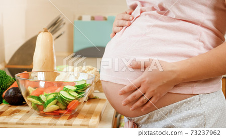 CLoseup of young pregnant woman cooking and eating vegetable salad holding big abdomen and touching it with hands CLoseup of young pregnant woman cooking and eating vegetable salad holding big abdomen and touching it with hands 73237962