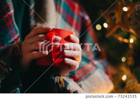Woman holding cozy red mug on Christmas tree background. Girl with hot drink - tea, coffee or cocoa Woman holding cozy red mug on Christmas tree background. Girl with hot drink - tea, coffee or cocoa 73241535