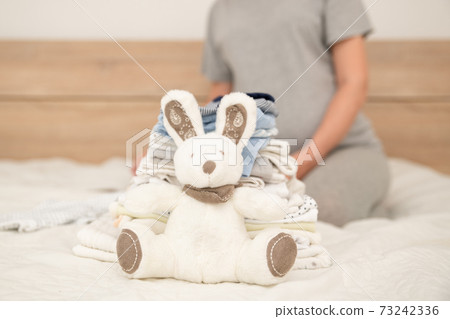 Pile of baby clothes, rabbit and pregnant woman on a bed Pile of baby clothes, rabbit and pregnant woman on a bed 73242336