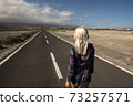 Blonde long hair girl viewed fromback walk on a long straight asphalt road - concept of freedom and independence - hope and future for next destination in life 73257571