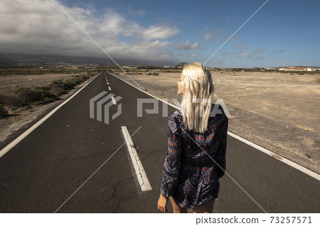 Blonde long hair girl viewed fromback walk on a long straight asphalt road - concept of freedom and independence - hope and future for next destination in life 73257571