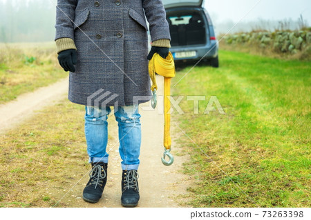Woman holding tow rope with hook near broken car on rural road 73263398