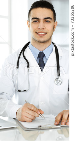Arab doctor man is using laptop computer while filling up medication history records form at the glass desk in r clinic. Medicine concept Arab doctor man is using laptop computer while filling up medication history records form at the glass desk in r clinic. Medicine concept 73265286