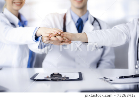 Group of doctors joining hands as a circle and ready to help patients while sitting at the desk in modern hospital office. Successful teamwork in medicine, Coronavirus countering concept Group of doctors joining hands as a circle and ready to help patients while sitting at the desk in modern hospital office. Successful teamwork in medicine, Coronavirus countering concept 73268412