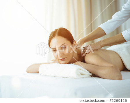 Pretty brunette woman enjoying procedure of back massage in sunny spa salon. Beauty concept. Cold toned picture Pretty brunette woman enjoying procedure of back massage in sunny spa salon. Beauty concept. Cold toned picture 73269126
