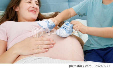 Little toddler boy playing with blue baby boots on big belly of young mother lying on bed. Concept of pregnancy, preparing and expecting child. Little toddler boy playing with blue baby boots on big belly of young mother lying on bed. Concept of pregnancy, preparing and expecting child. 73269275