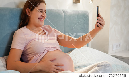 Smiling pregnant woman waiting for baby sitting in bed and calling friend or doctor on video call conference. Pregnant woman using smartphone 73270356