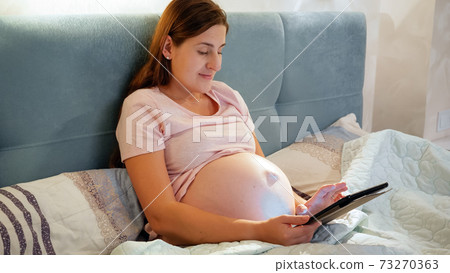 Smiling pregnant woman in pajamas using tablet computer and typing message while lying in bed at night 73270363