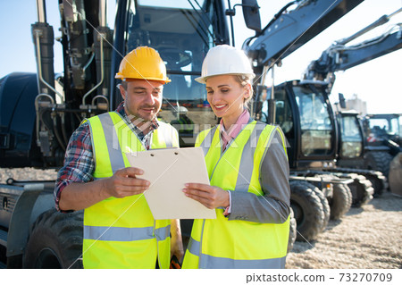 Two workers in open cast mine standing in front of heavy machinery 73270709