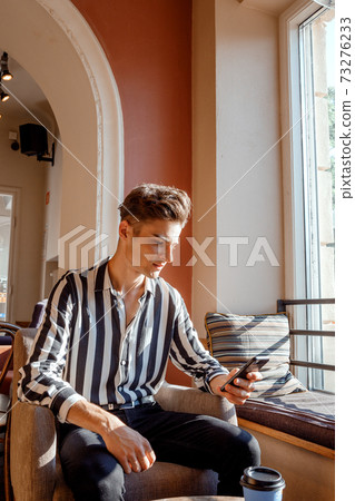 Young man sitting at a table in a cafe. Guy with a phone in a cafe on a chair. Stylish guy spends time with phone and coffee. 73276233