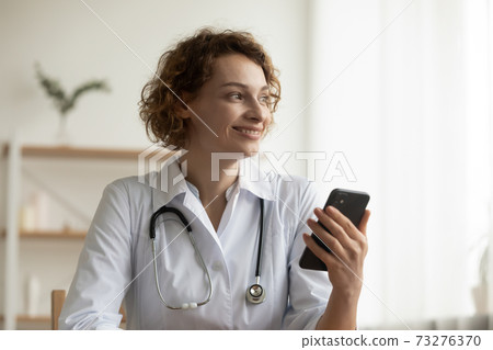 Happy dreamy young doctor looking in distance, holding mobile phone. 73276370