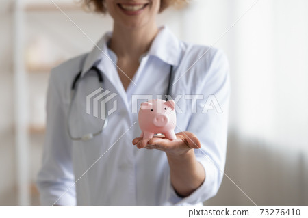 Close up smiling young doctor holding piggy bank in hands. Close up smiling young doctor holding piggy bank in hands. 73276410